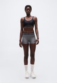 Αθλητικό άτομο όρθιο, φοράει μαύρο αθλητικό σουτιέν, γκρι σορτς Under Armour, λευκές κάλτσες και λευκά αθλητικά παπούτσια σε απλό φόντο.