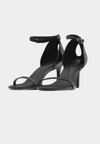 Paire de sandales noires à talons hauts avec fines brides à l’avant et lanières de cheville avec petites boucles dorées sur fond clair.