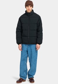 Zwarte pufferjack met hoge kraag, met gevoerde secties. Draag over lichtblauwe wijde jeans en donkerbruine schoenen. Bruine gebreide beanie op het hoofd.