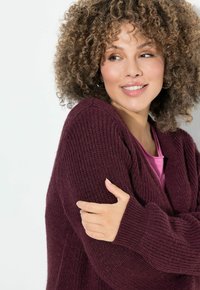 Cardigan in magenta lavorato a maglia con texture a coste, maniche lunghe e silhouette rilassata, indossato sopra una maglietta rosa. Aspetto morbido e caldo.