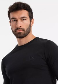 Černé tričko s dlouhým rukávem a kulatým výstřihem, se subtílně vyraženým logem poblíž ramene, vyrobené z hladkého a lehkého materiálu.