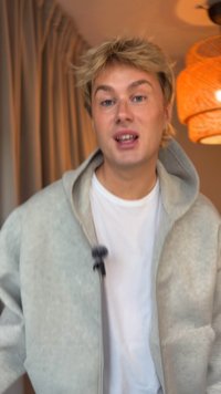 Felpa zip-up grigia chiara realizzata in materiale morbido, con un grande cappuccio, indossata sopra una maglietta bianca. Illuminazione soffusa con una lampada intrecciata sullo sfondo.