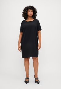 Robe noire à manches courtes avec une coupe décontractée, dotée d'un col rond. Associée à des escarpins noirs à bout pointu avec des brides décoratives.
