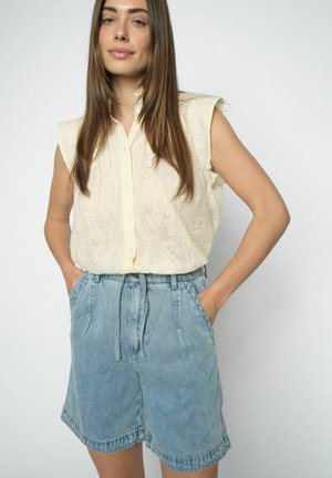 Woman with long brown hair wearing sleeveless cream blouse and high-waisted light blue denim shorts, hands in pockets, against white background.