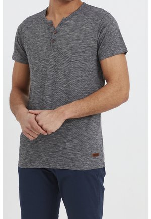 Mann trägt ein graues Henley-Shirt mit kurzen Ärmeln und drei Knöpfen sowie eine dunkelblaue Hose, die Hände vor dem Körper verschränkt vor weißem Hintergrund.
