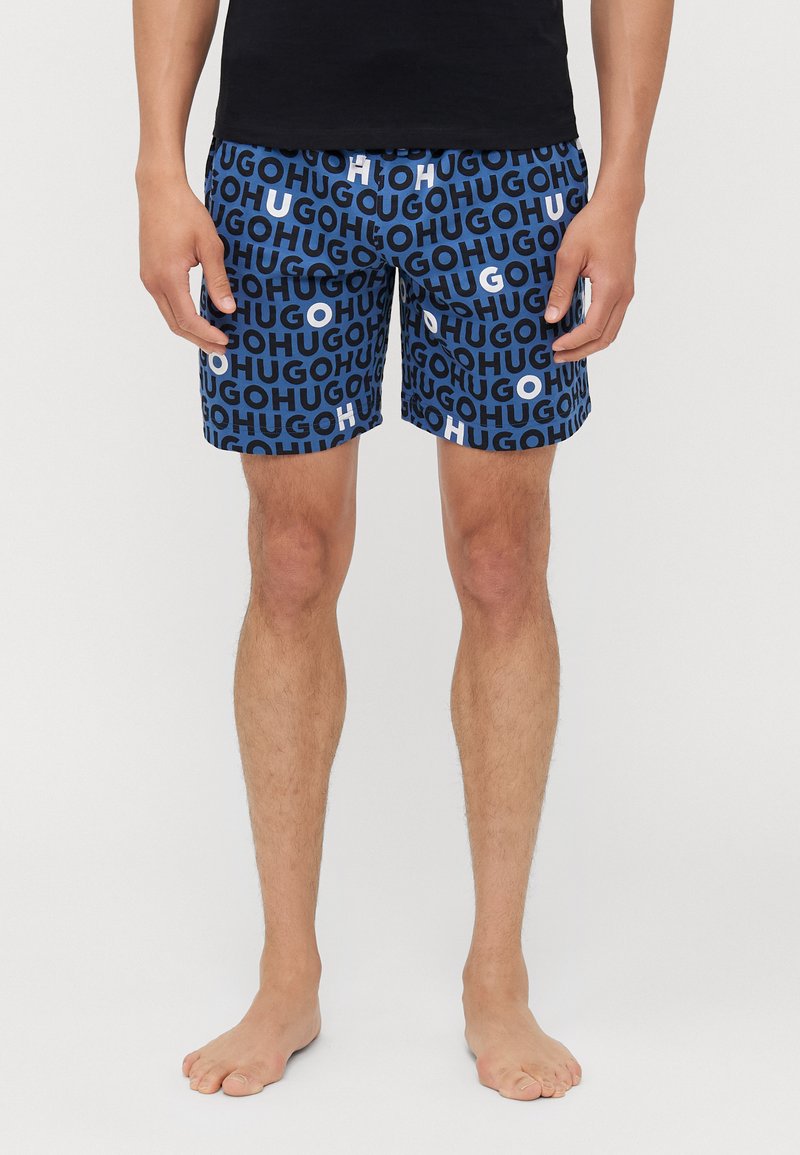 Shorts de bain bleu foncé, avec un motif de texte en gras répété en bleu clair et blanc ; taille élastique, coupe au niveau des genoux.