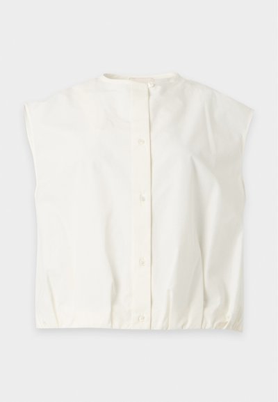 Blouse blanche à manches courtes avec col rond et fermeture par boutons sur le devant, étalée à plat sur un fond gris clair.