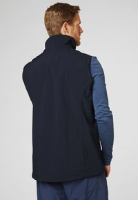 Marineblaues Softshell-Weste mit hohem Kragen, glatter Textur, ohne Verschlüsse und Schulternähten. Über einem blauen Langarmshirt getragen.