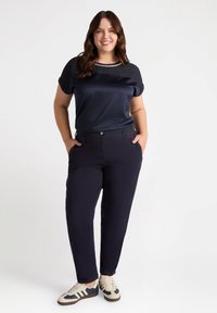 Top din satin navy cu mâneci răsucite, asortat cu pantaloni închiși la culoare, prevăzuți cu buzunare laterale. Încălțăminte casual în bej și navy cu dungi.