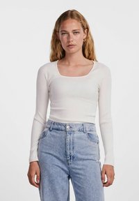 Top bianco a maniche lunghe con scollatura quadrata, abbinato a jeans azzurri a vita alta. Il tessuto sembra morbido e testurizzato.