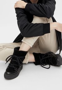 Sneaker alte nere con una texture pelosa alla caviglia, dotate di lacci neri spessi e suola in gomma nera, abbinate a pantaloni beige.