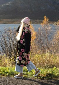 Dlhý čierny kvetinový kabát s ružovými a zelenými vzormi, nosený cez svetlomodré džínsy a béžové tenisky, doplnený ružovou čiapkou.