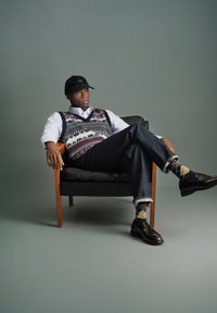 Man som bär mönstrad väst, vit skjorta, jeans, rutmönstrade strumpor, loafers och svart keps, sittande med korsade ben på en träram.