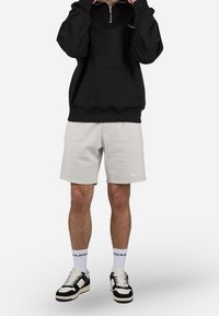 Sweat-shirt noir à col zippé, shorts gris clair, chaussettes blanches avec logo, baskets noires et blanches. Coupe confortable avec un design décontracté.