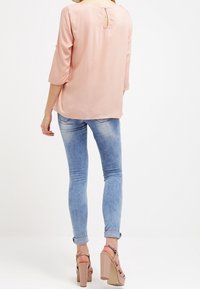 Blusa di seta leggera rosa chiaro con maniche a tre quarti e dettaglio a chiave sulla schiena, abbinata a jeans azzurri aderenti e sandali con tacco a blocco beige.
