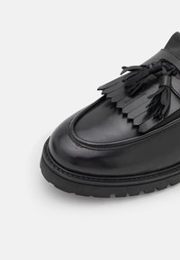 Chaussure en cuir noir avec un bout arrondi, semelle plate et des franges décoratives sur le devant. Fini brillant avec des coutures renforcées.