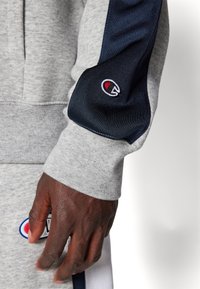 Sudadera gris con detalles en azul marino, puños acanalados y un pequeño logotipo bordado en la manga. Presenta una tela suave y texturizada.