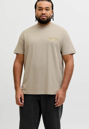 Homme aux cheveux tressés et barbe portant un T-shirt beige avec un petit texte décoratif sur la poitrine et un pantalon noir, debout devant un fond uni.