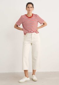 Raudonos ir baltos dryžuotos trumpomis rankovėmis marškiniai derinami su aukštu liemeniu, kreminės baltos spalvos plačiomis kelnėmis ir baltomis sportbačių, stovinčiais ant neutralios fono.