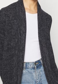 Cardigan grigio scuro con una texture morbida, davanti aperto e fit rilassato. Indossato sopra una camicia bianca, abbinato a jeans azzurri chiari.