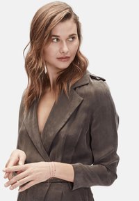 Veste ajustée à col V plongeant en tissu gris doux et texturé, avec des manches longues et des épaulettes. Le mannequin porte un bracelet délicat au poignet.