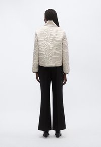 Sieviete ar gariem bizēm, ģērbusies gaišā jostiņā ar abstraktām līnijām, melnās platām biksēm un melniem apaviem, stāv ar muguru pret skatītāju.