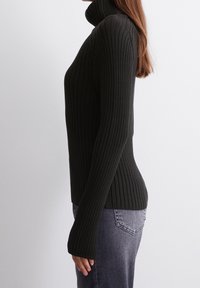 Pull à col roulé noir côtelé avec manches longues, au design ajusté et à la texture douce. Associé à un jean gris, mettant en valeur une silhouette élégante.