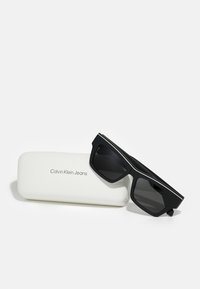 Gafas de sol negras rectangulares con un marco grueso, presentadas junto a un estuche blanco grabado con "Calvin Klein Jeans."