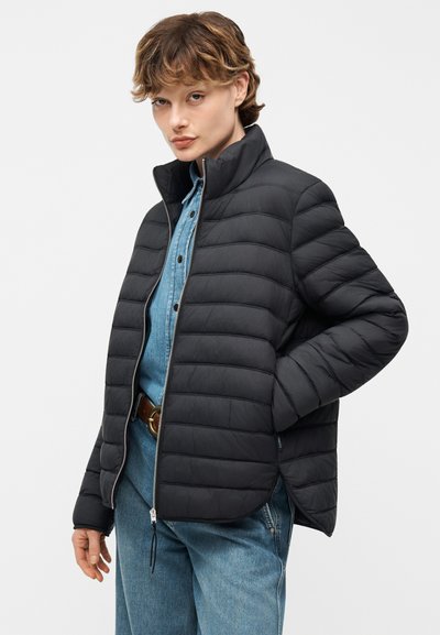 Νεαρός με κοντά μαλλιά που φοράει μαύρο καπιτονέ μπουφάν πάνω από ένα κουμπωτό τζιν πουκάμισο και μπλε τζιν, στέκεται με το ένα χέρι στην τσέπη.