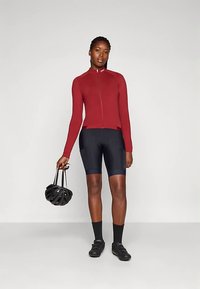 Tenue de cyclisme comprenant un maillot rouge ajusté à manches longues, un short noir et un casque noir tenu dans la main gauche. Tissu texturé visible.