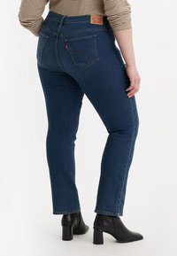 Tmavě modré džíny z denimu s přiléhavým střihem, s zadními kapsami a koženou nášivkou značky. Nošeny s černými kotníkovými botami.