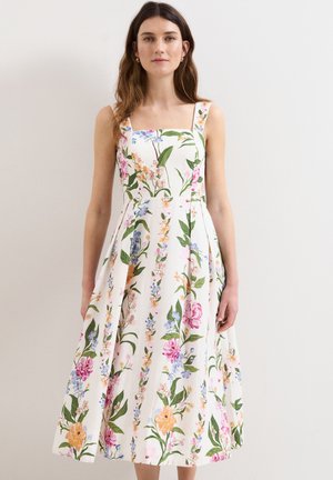 Vrouw die een witte mouwloze midi-jurk draagt met roze, oranje, blauwe en groene bloemmotieven, staand tegen een effen lichte achtergrond.
