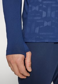 Langarm navy atletisch shirt met duimopeningen, met een subtiel patroon en zachte, stretchy stof. Close-up van de mouw en hand.
