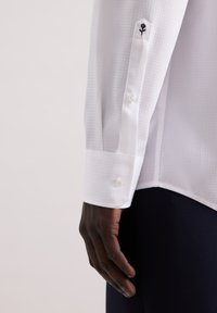 Witte lange mouw shirt van een gestructureerde stof, met een knoopsluiting bij de manchetten en een geborduurd bloemenmotief nabij de manchetten.