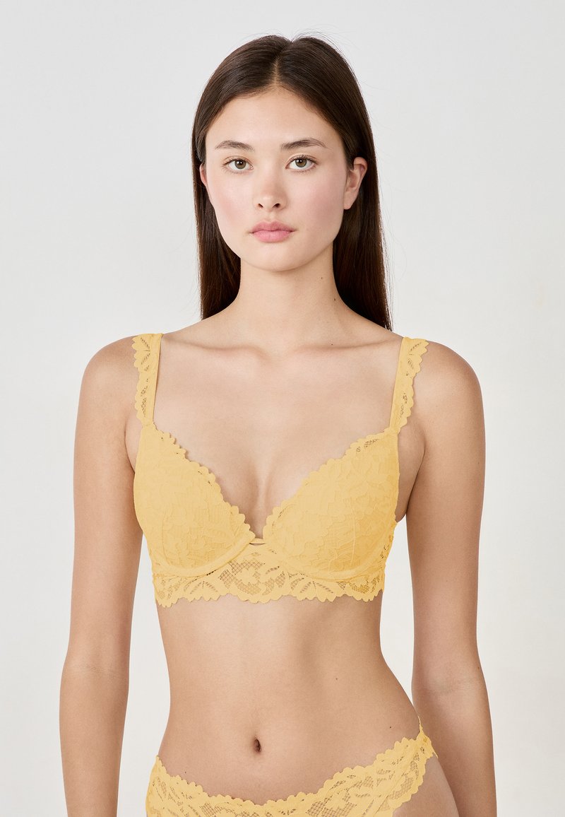 Soutien-gorge en dentelle jaune avec des bords festonnés, un décolleté plongeant et des bretelles ajustables. Le tissu présente un motif floral et une finition texturée.