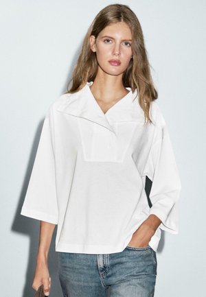 White oversized shirt with a wide collar, short sleeves, and a subtle front pleat. Worn with light blue jeans. Soft cotton fabric.