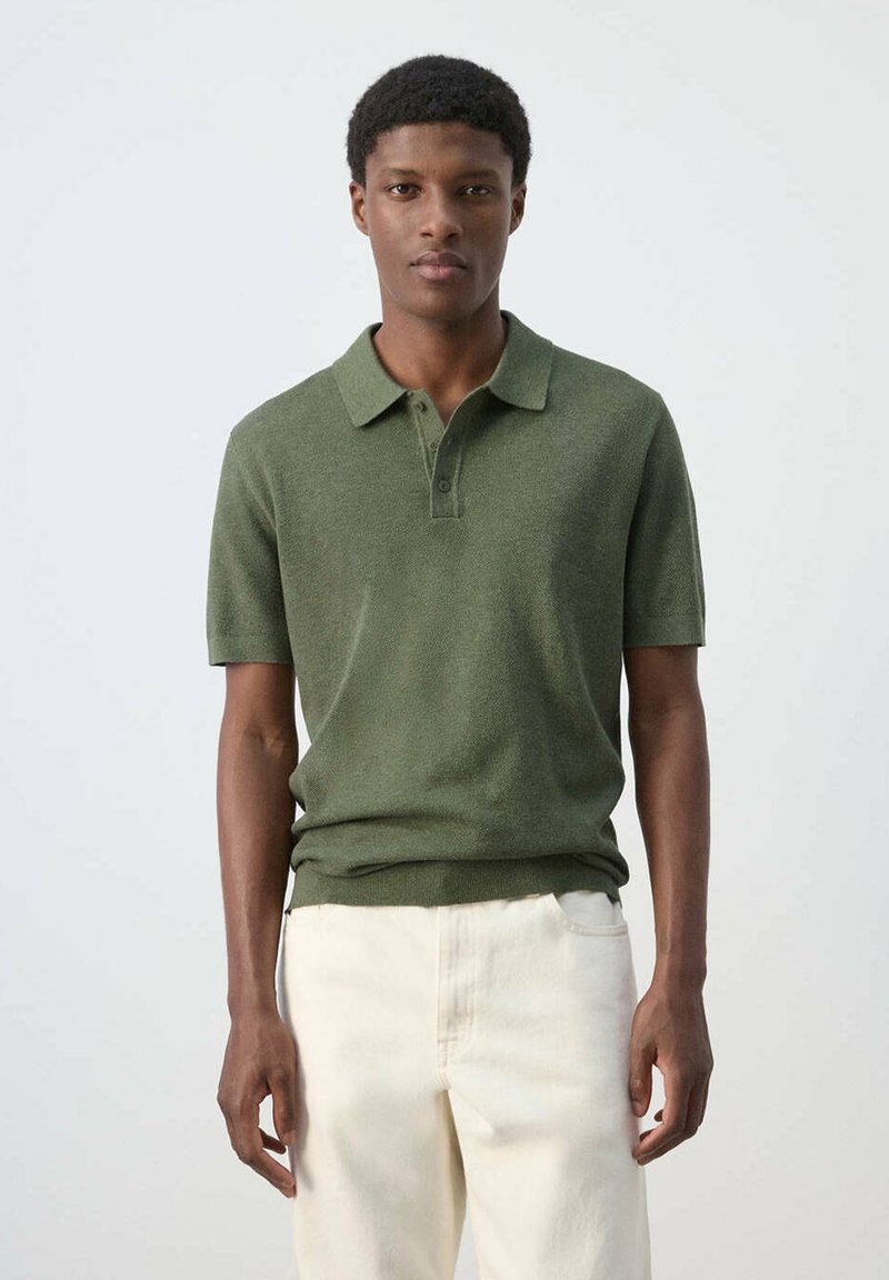 Jeune homme debout portant un polo vert à manches courtes et un pantalon beige clair sur un fond blanc uni.