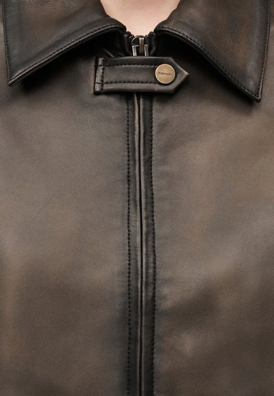 Κοντινό πλάνο για το γιακά σκούρας δερμάτινης μπουφάν με φερμουάρ και λουράκι που διαθέτει στρογγυλό μεταλλικό κουμπί με χαραγμένο το όνομα "Holzweiler".