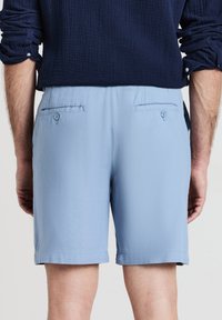 Hellblaue Shorts aus strukturiertem Stoff, mit zwei Gesäßtaschen mit Knöpfen und einem geraden Saum. Getragen mit einem marineblauen Hemd.