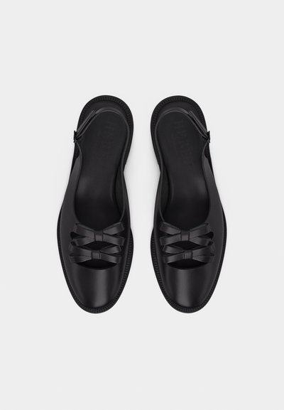 Μαύρα δερμάτινα φλατ σανδάλια με διπλά λουριά φιόγκου πάνω από τα δάχτυλα και λουράκι αστραγάλου με λουρί πίσω, παρουσιασμένα από πάνω σε λευκό φόντο.