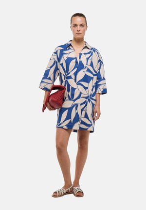 Femme portant une robe-chemise à motif feuilles bleu et beige, tenant un sac tissé rouge et des sandales plates blanches.