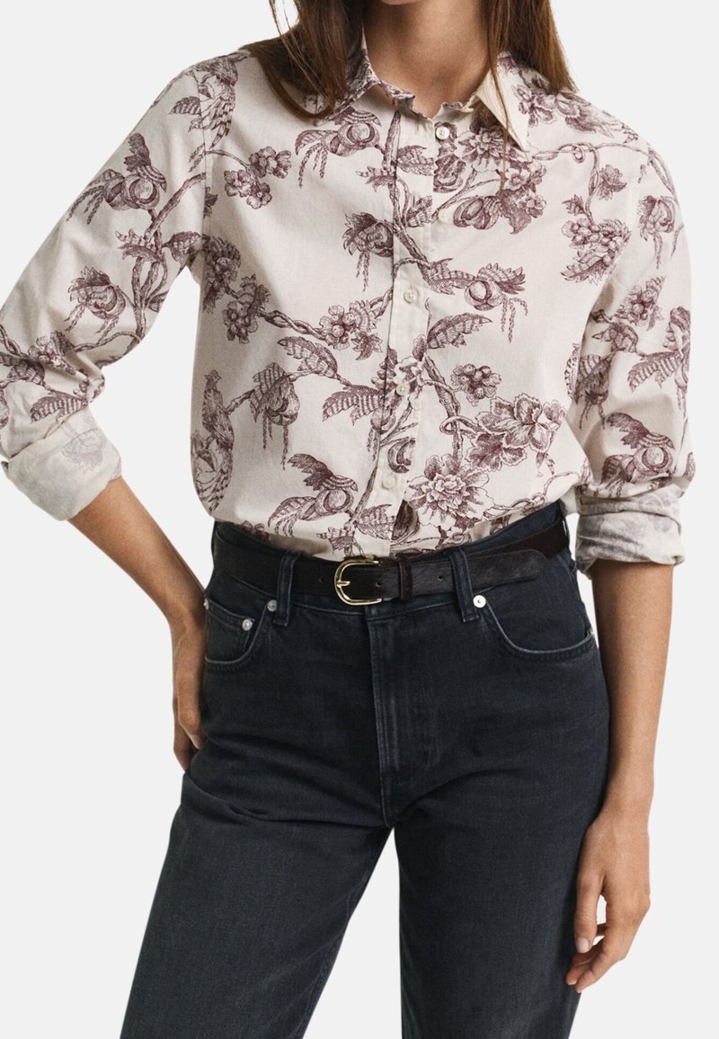 Femme portant une chemise beige à boutons avec un motif floral bordeaux, manches retroussées, rentrée dans un jean noir taille haute avec ceinture noire.