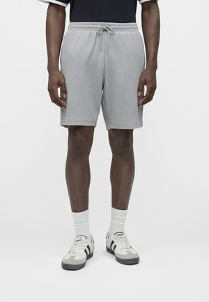 Homme portant un short côtelé gris clair avec cordon, des chaussettes blanches mi-mollet et des baskets beige avec des rayures noires, debout devant un fond uni.