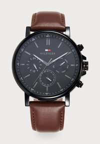 Reloj cronógrafo negro con esfera negra, agujas plateadas y correa de cuero marrón. Incluye indicadores de fecha y día, con marcadores de hora plateados.