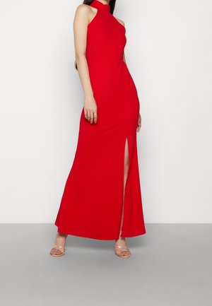Kobieta ubrana w czerwoną, bezrękawnikową suknię maxi z wysokim kołnierzem i bocznym rozcięciem, w połączeniu z sandałkami na obcasie z odkrytymi palcami na jednolitym tle.