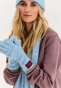 Světle modré rukavice z měkkého, texturovaného materiálu s bordó štítkem. Modrý šál sladěný s rukavicemi je omotaný kolem krku.