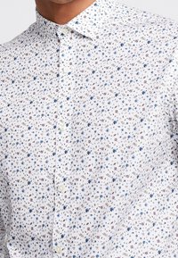 Biała koszula z małym kwiatowym wzorem w niebieskim i czerwonym kolorze, z kołnierzykiem na guziki i lekką teksturą, idealna na codzienne noszenie.