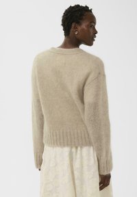 Beiger gestrickter Pullover mit weicher, flauschiger Textur, mit langen Ärmeln und einem gerippten Saum, getragen über einem cremefarbenen, mit Blumen bedruckten Rock.