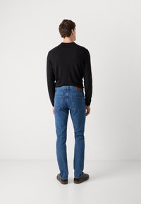 Camisa negra de manga larga, vaqueros azules con un parche de cuero marrón, diseño ajustado, textura suave, bolsillos traseros visibles, calzado casual.