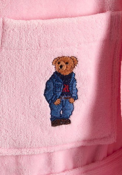 Ours en peluche brun brodé portant une veste en jean bleue, un jean et des chaussures marron sur un tissu rose doux texturé.